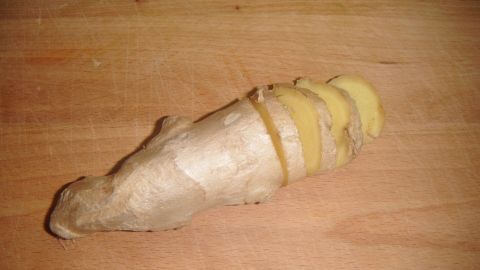 Ein Stück Ingwer liegt auf einem Holzbrett, es wird in dünne Scheiben geschnitten, um ihn einfacher zu schälen.