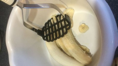 Eine Gabel zerdrückt eine Banane in einer weißen Schüssel auf einer Arbeitsfläche, um Banane für ein Rezept vorzubereiten.