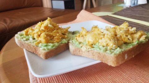 Zwei Scheiben Vollkornbrot mit Avocado und Rührei liegen auf einem weißen Teller, umgeben von einem hellem Wohnraum.