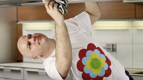Ein Mann wischt Fett und Schmiere von einem Küchenschrank, während er näher an die Decke heranreicht. Die Küche ist modern eingerichtet.