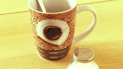 Ein Kaffeebecher steht auf einem Tisch, mit einem Löffel darin; daneben befindet sich ein Zuckergefäß. Ränder an Kaffeebecher entfernen.