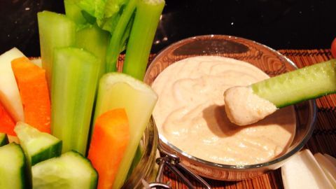 In einer Schale mit Erdnuss-Cashew-Kokos-Dip wird ein Selleriestick eingetaucht, umgeben von bunten Gemüsesticks.