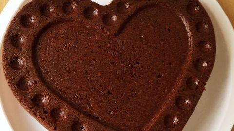 Ein herzförmiger Schoggikuchen liegt auf einem weißen Teller, umgeben von einer Holzoberfläche, bereit zum Servieren.