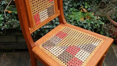 Ein Kaffeehausstuhl mit geflochtenem Sitz, der mit Kreuzstich bestickt wird, steht in einem begrünten Außenbereich.