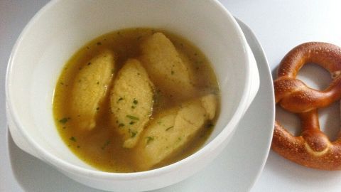 Eine weiße Schale mit Grießnockerlsuppe auf einem Teller, daneben liegt eine Brezel auf einem weißen Tisch.