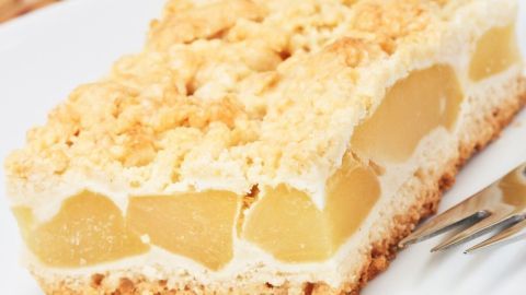 Ein saftiges Stück Apfelkuchen aus Hefeteig mit Streuseln liegt auf einem weißen Teller, umgeben von einem geflochtenen Untergrund.