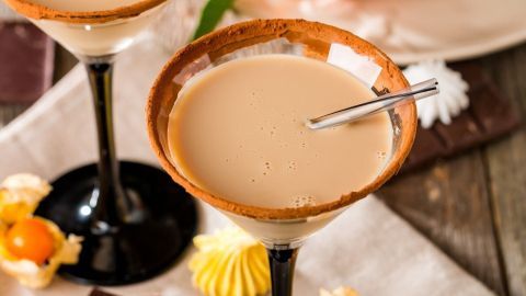 Zwei elegante Cocktails mit einer cremigen, bernsteinfarbenen Flüssigkeit stehen auf einem rustikalen Tisch, umgeben von Früchten.