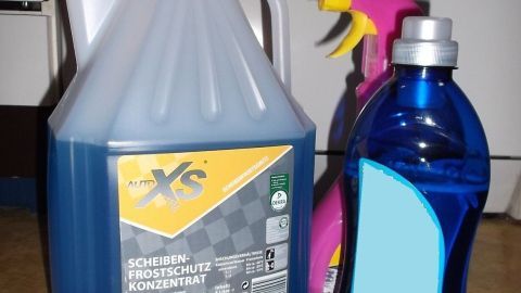 Ein blauer Kanister für Wasser und zwei Sprayflaschen stehen auf einem Tisch in einer Garage oder Werkstatt, bereit für die Scheibenwaschanlage.