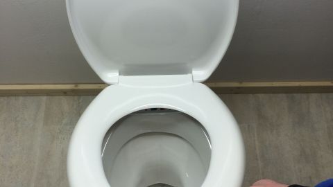Eine Hand greift nach einem Toilettensitz, um ihn herunterzuklappen. Die Umgebung ist einfach und modern gestaltet.