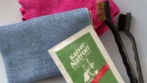 Ein Reinigungstuch und eine Packung "Kaiser Natron" liegen neben zwei Bürsten auf einem Tisch, bereit zur Reinigung dunkler Fugen von Bodenfliesen.