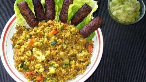 Kleine, gebratene Hackbällchen liegen auf einem bunten Curry-Gemüsebratreis, umgeben von frischem Salat und einer Schüssel Beilage.