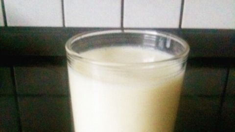 Ein Glas Bananen-Buttermilch steht auf einer dunklen Fläche, umgeben von einer gefliesten Wand im Hintergrund.