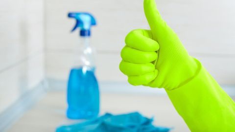 Eine grüne Gummihand zeigt Daumen hoch, während ein Reinigungsmittel und ein Tuch auf lackierten Fliesen im Hintergrund stehen.