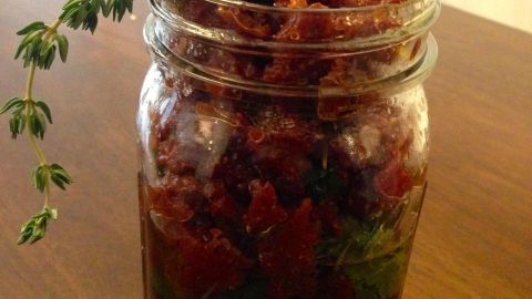 Ein Glas mit getrockneten Tomaten, gefüllt mit frischen Kräutern, steht auf einem Holztisch und strahlt Genuss aus.