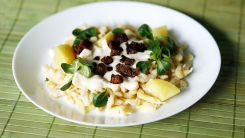 Spätzle und Kartoffeln liegen in einer Schüssel, bedeckt mit Käsesoße und frischen Kräutern, auf einem grünen Tisch.