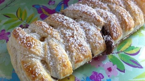 Ein Blätterteigstrudel liegt auf einem bunten Tisch, überzogen mit Puderzucker und gefüllt mit Schokolade.