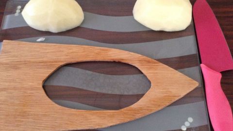 Auf einem Holzbrett liegt eine Schneidehilfe für Fächerkartoffeln, während zwei geschälte Kartoffeln und ein Messer neben ihr platziert sind.