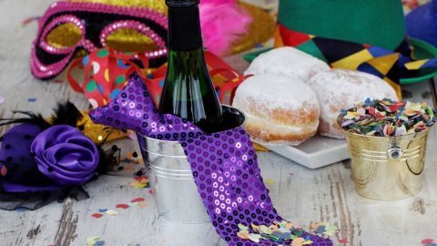 Eine lila Glitzer-Hochschuhhälfte hält eine Flasche Sekt, umgeben von bunten Karnevalsaccessoires und Berliner Pfannkuchen.
