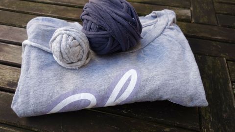 Ein graues T-Shirt liegt gefaltet auf einem Holztisch, während zwei Knäuel Garn aus alten T-Shirts daneben liegen.