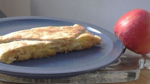 Ein Stück Apfelkuchen mit Dinkelgrieß liegt auf einem blauen Teller, während ein roter Apfel daneben ruht.