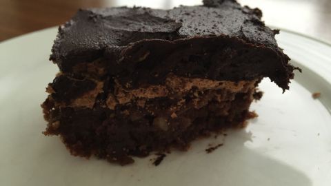Ein Stück Schoko-Brownie liegt auf einem weißen Teller, dunkel und saftig, umgeben von natürlichem Holzlicht.