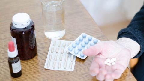 Eine Hand hält mehrere Tabletten, während ein Glas Wasser und verschiedene Fläschchen auf einem Tisch liegen.