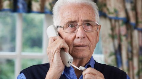 Ein älterer Mann spricht besorgt am Telefon. Im Hintergrund sind Blumenvorhänge und Tageslicht sichtbar.