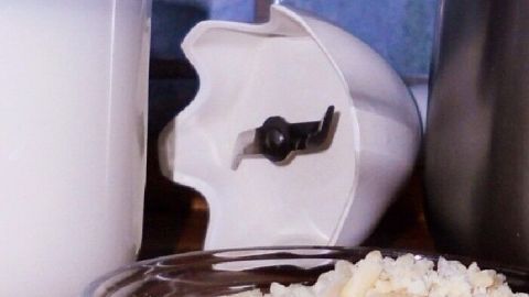 Ein Glas Mandel-Reismilch steht neben einem Mixbehälter und einer Schüssel mit geriebenem Reis auf einer Holzoberfläche.