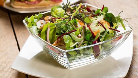 Ein bunter Salat aus frischem Gemüse und Kräutern steht in einer Glas-Schüssel auf einem weißen Teller, umgeben von Holz.