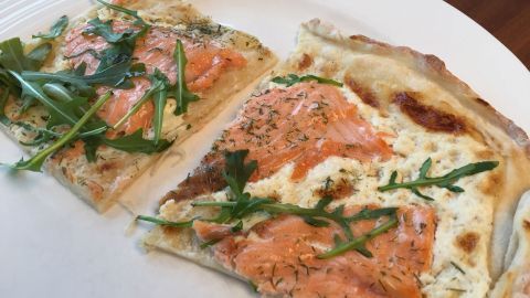 Flammkuchen mit frischem Lachs und Rucola liegt auf einem weißen Teller, die Stücke sind leicht angehoben und dekoriert.