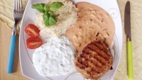 Ein saftiges griechisches Hacksteak liegt auf einem Teller, ergänzt durch Reis, Tomaten und eine Joghurtsoße im Hintergrund.