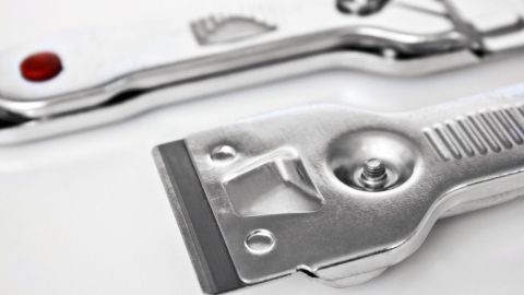 Ein Metallkratzer liegt auf einer glatten Oberfläche, bereit, um Farbflecken und Klebereste vom Fenster zu entfernen.