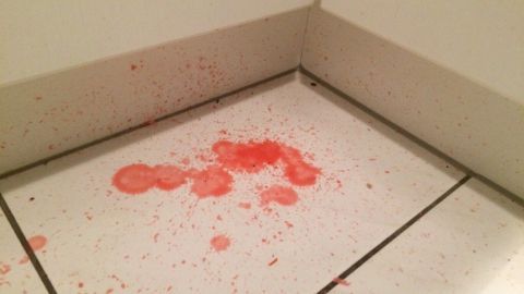 Dunkelrotes Kerzenwachs ist auf weißen Fliesen verschüttet, mit Tropfen und Spritzern überall im Eckenbereich verteilt.