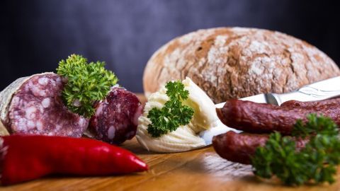 Ein rustikales Brot liegt neben Salami und Käse, während frische Kräuter und rote Paprika die Szene ergänzen.