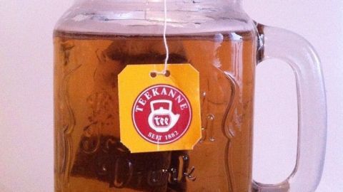 Ein Teebeutel hängt in einem Glasbecher mit warmem Tee, während die Becher auf einem hellen Tisch steht.