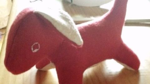 Ein roter, genähter Hund mit einem weißen Ohr steht auf einem Holztisch, umgeben von Fäden und Nähutensilien.