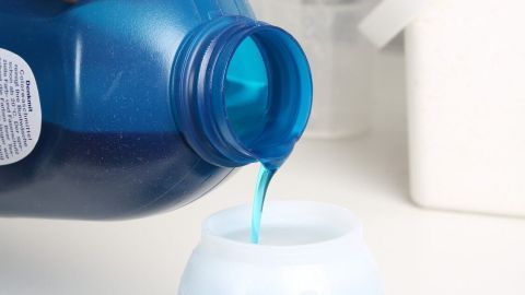 Eine Hand gießt blauen Duft in eine kleine, runde Schale in einer hellen Badezimmerumgebung, um frischen Duft zu erzeugen.