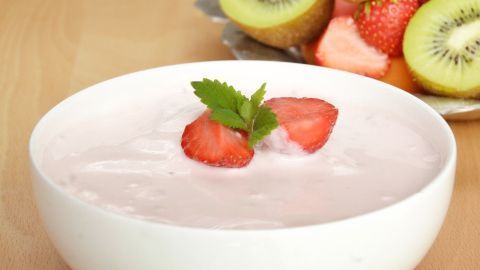 Eine Schüssel Joghurt mit Erdbeeren und Minze oben drauf steht auf einem Tisch, umgeben von frischem Obst.