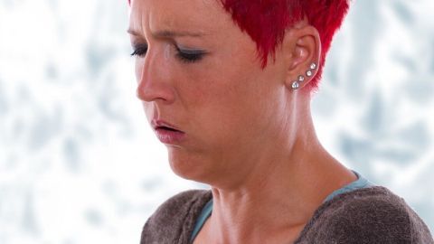 Eine Frau mit rotem Haar hält sich den Brustkorb und zeigt Anzeichen von Husten, angedeutet durch ihr angespanntes Gesicht.
