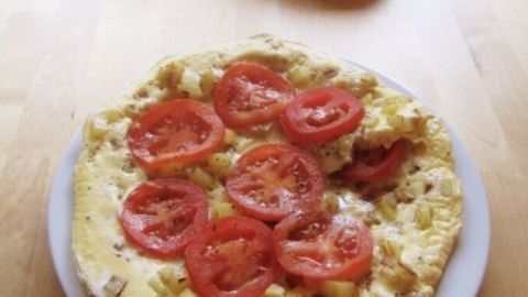 Ein Kartoffel-Omelett liegt auf einem Teller, belegt mit frischen Tomatenscheiben, auf einem Holztisch in einer hellen Küche.