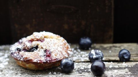 Gesunde Muffins mit Heidelbeeren liegen auf einem rustikalen Tisch, umgeben von frischen Beeren und Puderzucker.