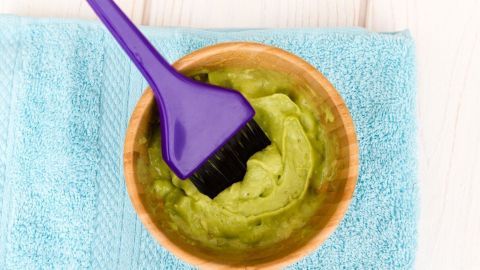 Eine lila Bürste rührt eine grüne Avocado-Maske in einer Schüssel, um trockene Haut zu pflegen, auf einem blauen Tuch.