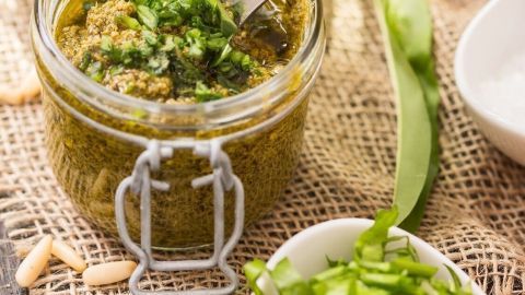 Ein Glas Bärlauchpesto steht auf einem rustikalen Tisch, während frischer Bärlauch und gehackte Kräuter daneben liegen.