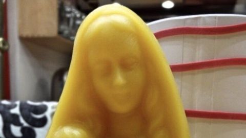 Eine gelbe Wachsfigur zeigt eine Mutter mit einem Kind, die auf einem Tisch steht, umgeben von einer gemütlichen Umgebung.