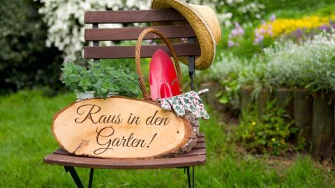 Ein Holzschild mit der Aufschrift "Raus in den Garten!" steht auf einem Stuhl, umgeben von Pflanzen und einer Sommermütze.