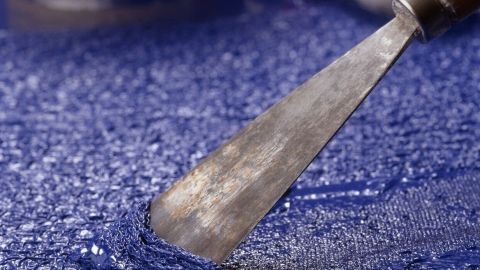Ein Spachtel bewegt sich über eine blaue Oberfläche, während ein Abbeizer im Hintergrund steht.