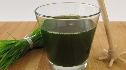 Ein Glas mit grünem Gerstengrassaft steht auf einem Holztisch, während ein Bündel frisches Gras neben ihm liegt.