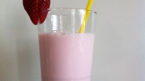 Ein Glas Erdbeermilch steht auf einem grünen Untergrund, mit einem Strohhalm und einer Erdbeere am Rand, cremig und rosa.