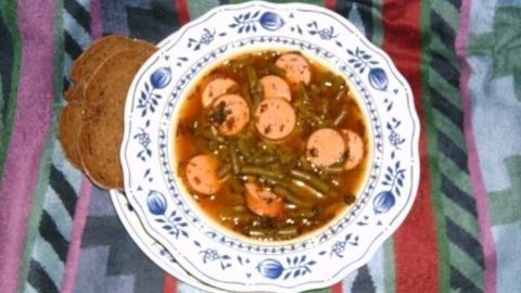 Eine Schüssel Bohnensuppe mit Bockwurststückchen liegt auf einem bunten, gestreiften Tuch, umgeben von einem rustikalen Teller.