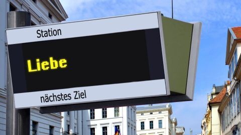 Eine Wegweisung zeigt „Liebe“ als Ziel an; eine attraktive urbane Umgebung unter blauem Himmel umgebt die Station.